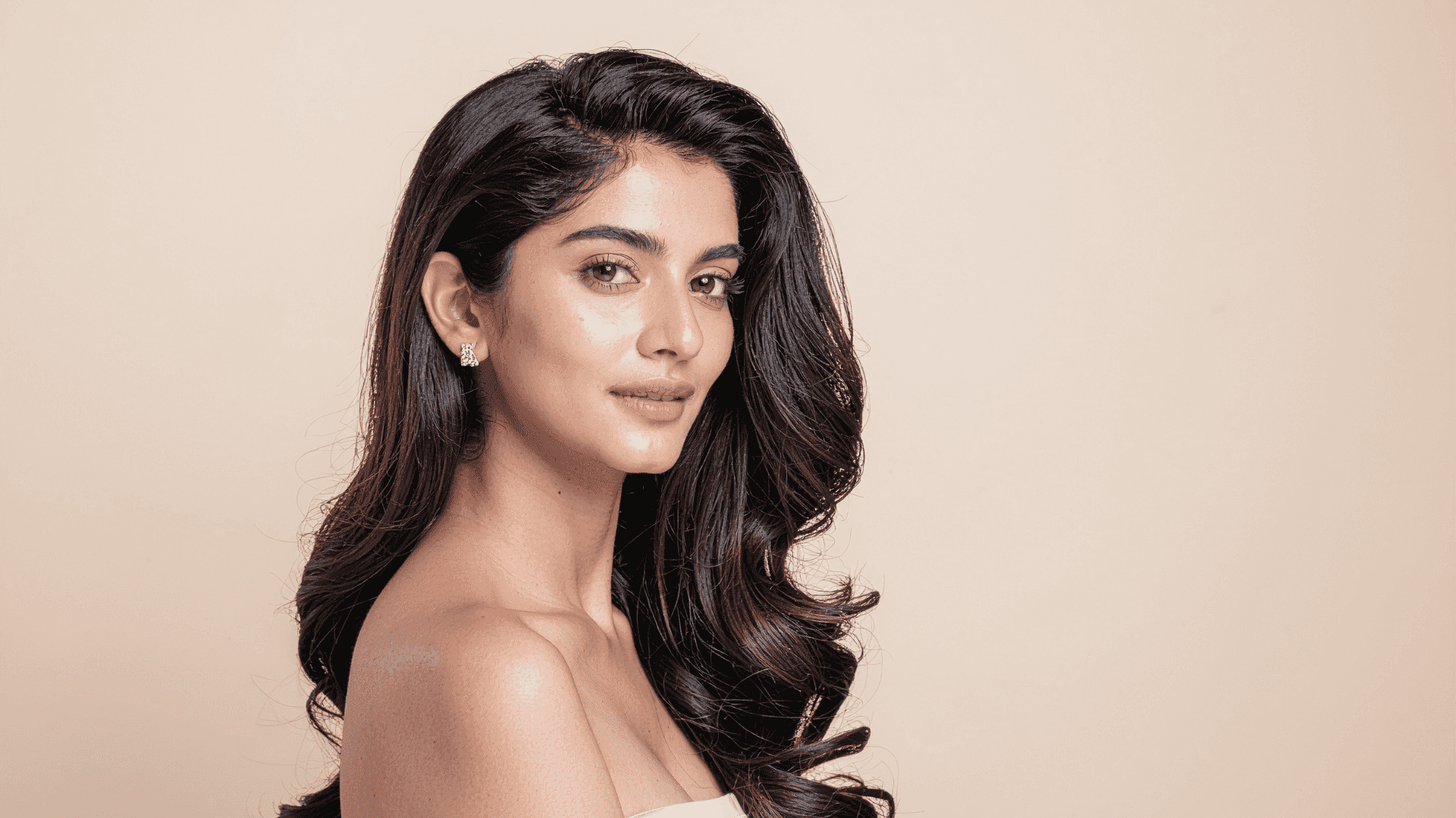 Woman with long dark hair and elegant earrings in portrait against a beige background.