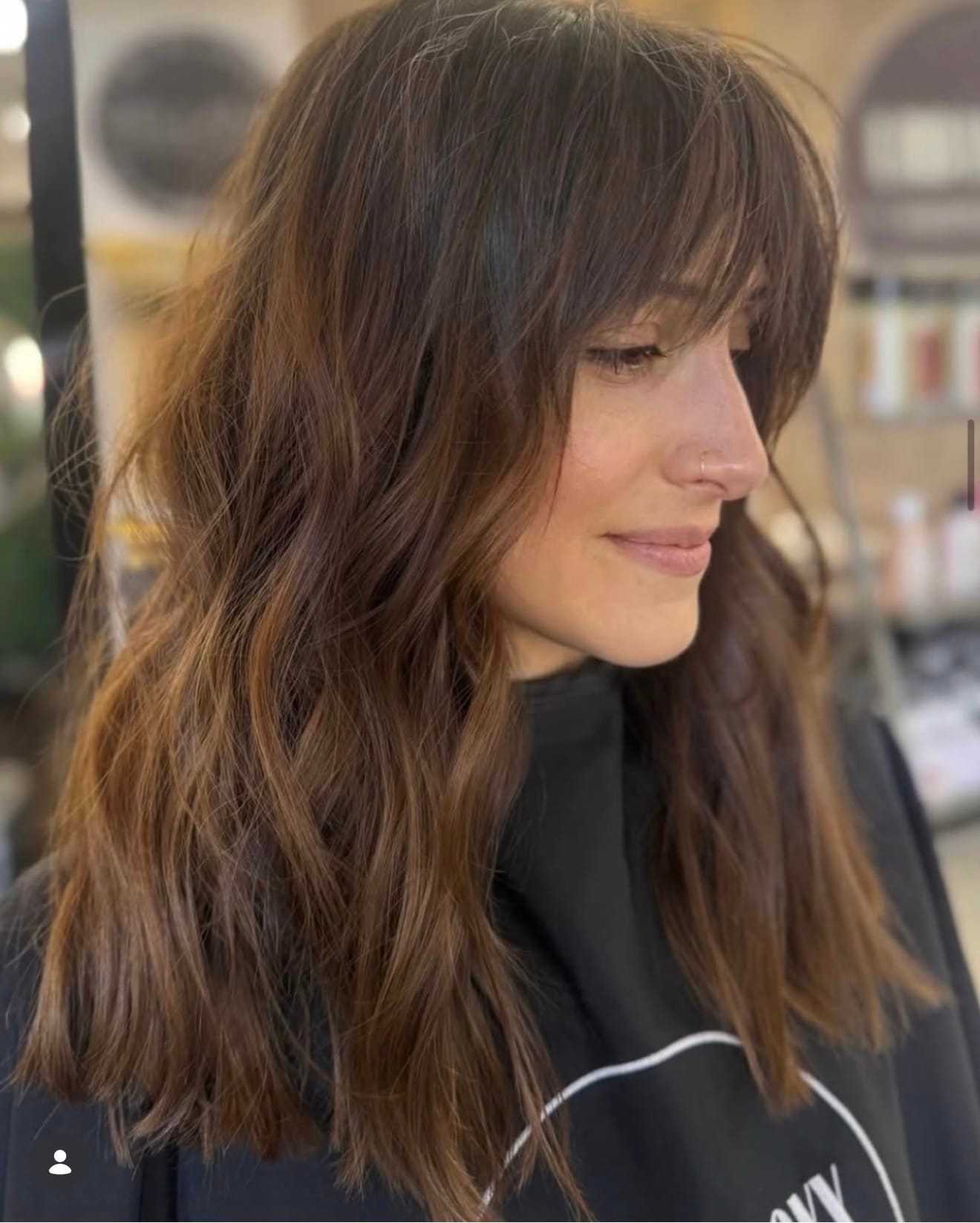 Woman with wavy brown hair and bangs at a salon, wearing a black cape.